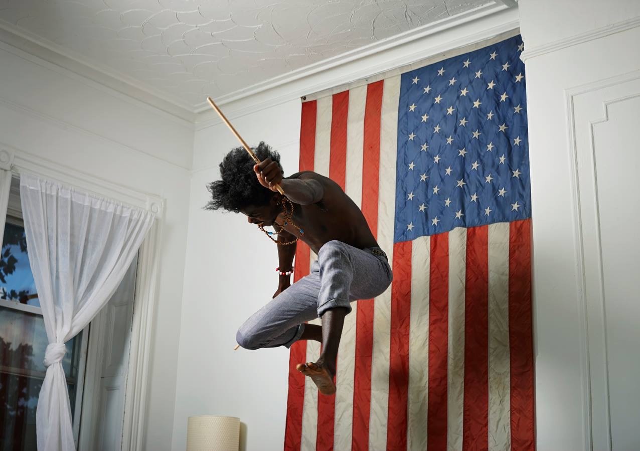Pauli Lovejoy suspended mid-air, drumstick raised, against a hanging flag.