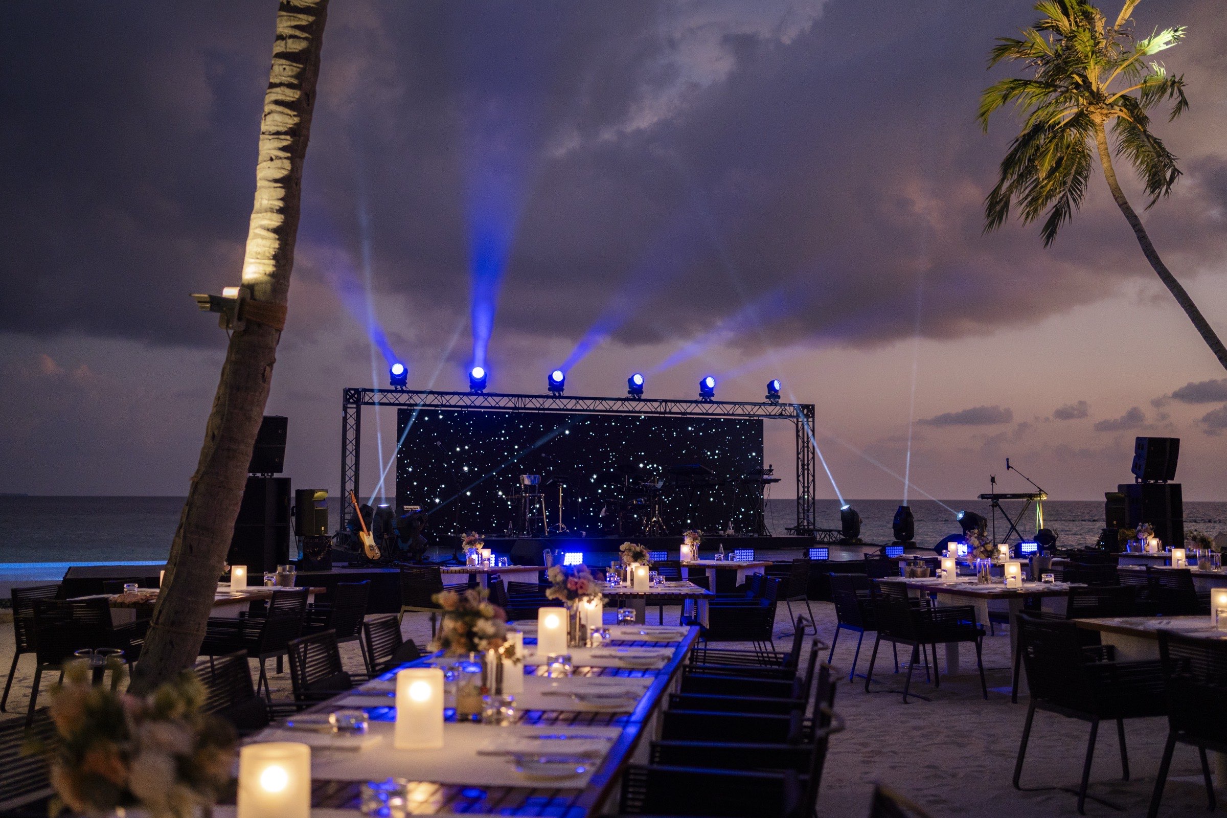 A stage lit with blue beams on the beach at dusk, candlelit tables set in the sand in front of it.
